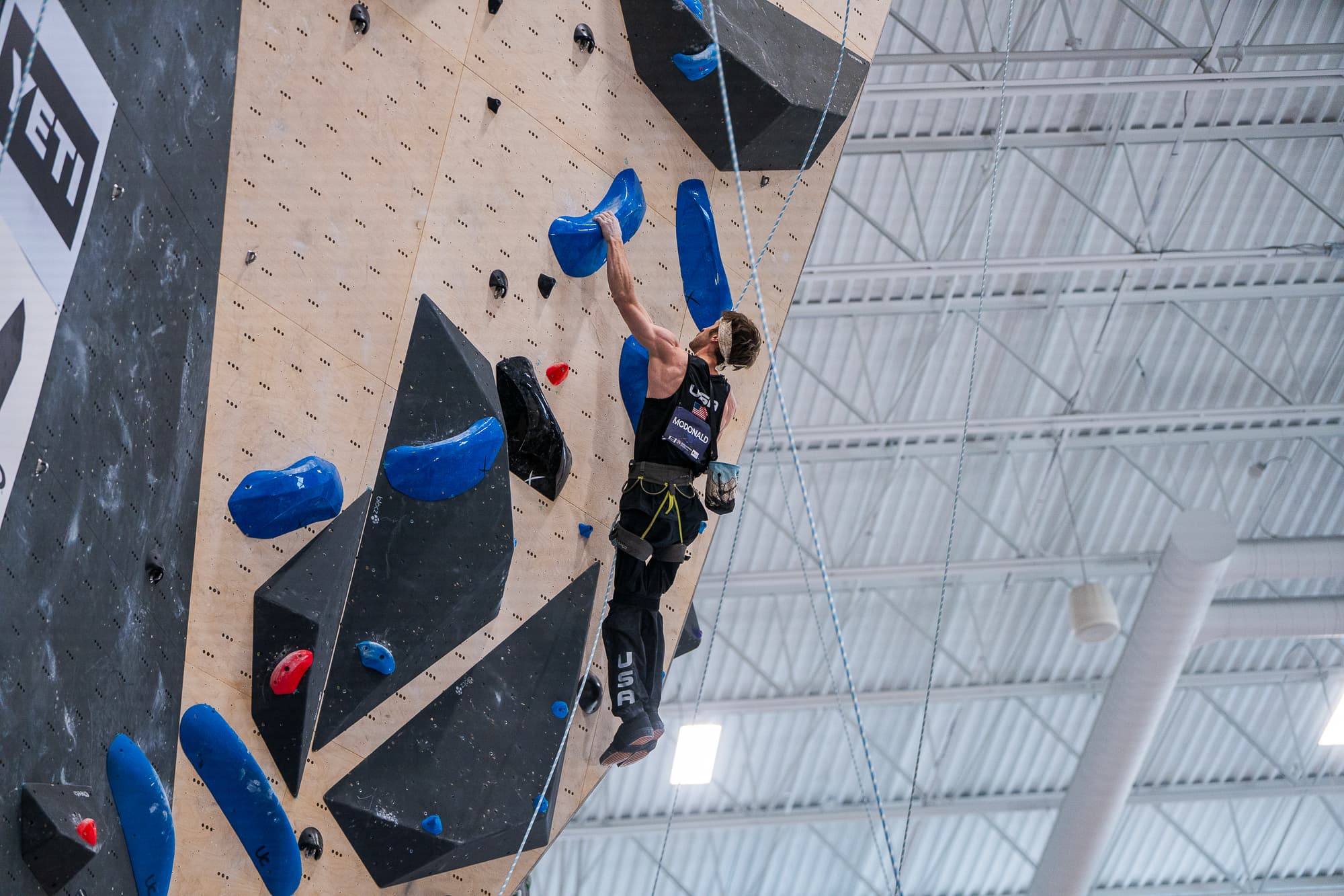 Caleb McDonald mid-route on a steep competition wall in his USA MCDONALD singlet
